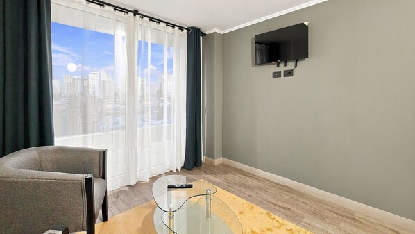 A room with a city view, a flat-screen TV mounted on the wall, a glass coffee table, and a chair.