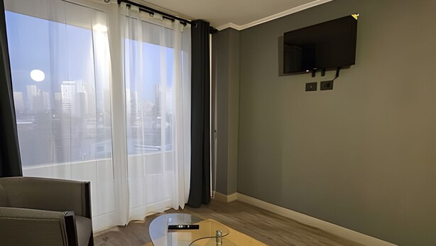 Habitació moderna amb taula de vidre, televisió de pantalla plana penjada a la paret i vista a un paisatge urbà a través de grans finestres amb cortines lleugeres.