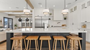 Cuina moderna amb una illa central, tamboretes de bar, electrodomèstics d'acer inoxidable i il·luminació penjant.