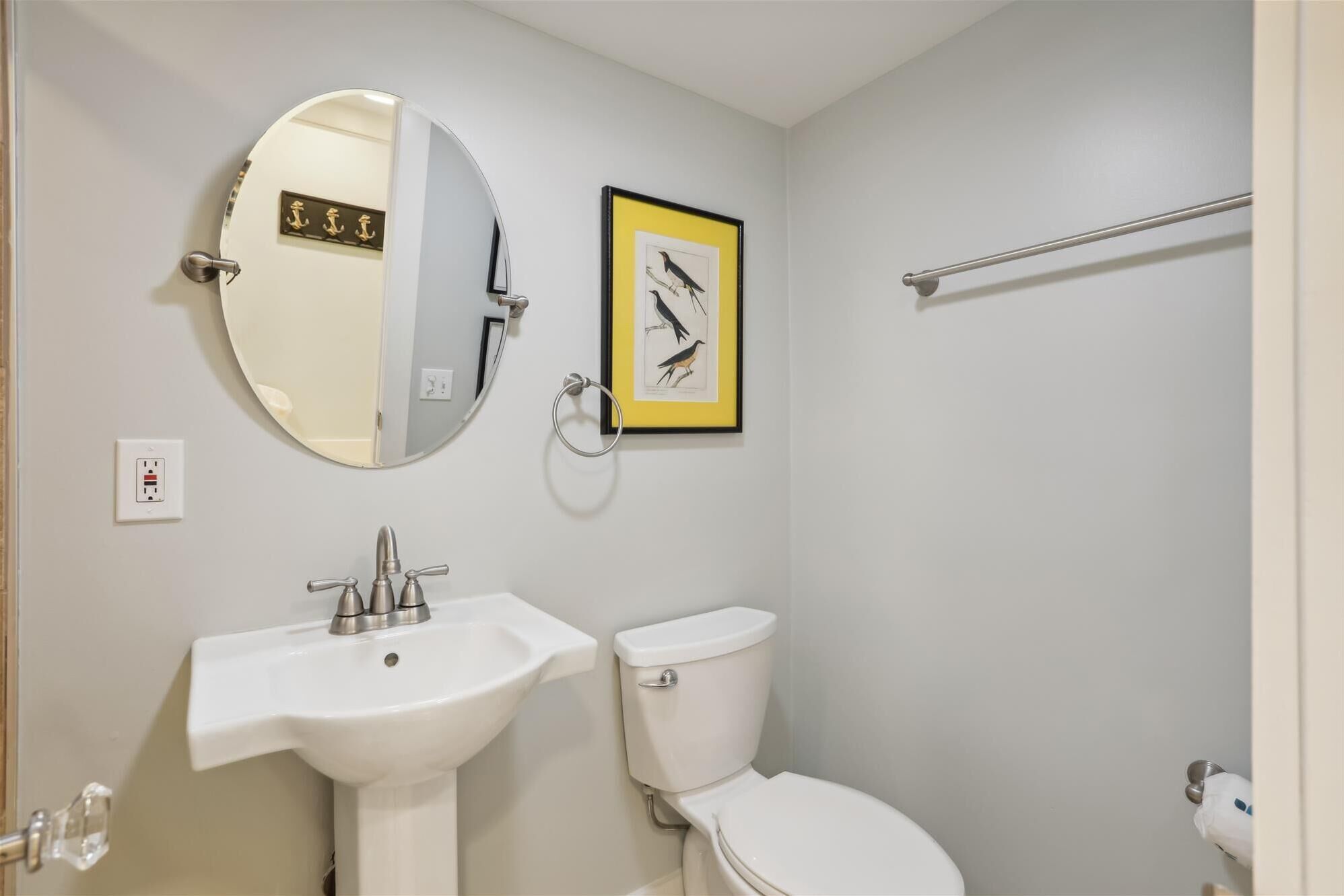Un baño con un lavamanos blanco, un inodoro, un espejo redondo y una ilustración enmarcada de un pájaro en la pared.