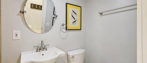 Un baño con un lavamanos blanco, un inodoro, un espejo redondo y una ilustración enmarcada de un pájaro en la pared.