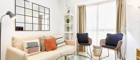 Una sala de estar con un sofá, dos sillones, una mesa de centro de vidrio, una lámpara de pie, una estantería y un amplio ventanal con cortinas.