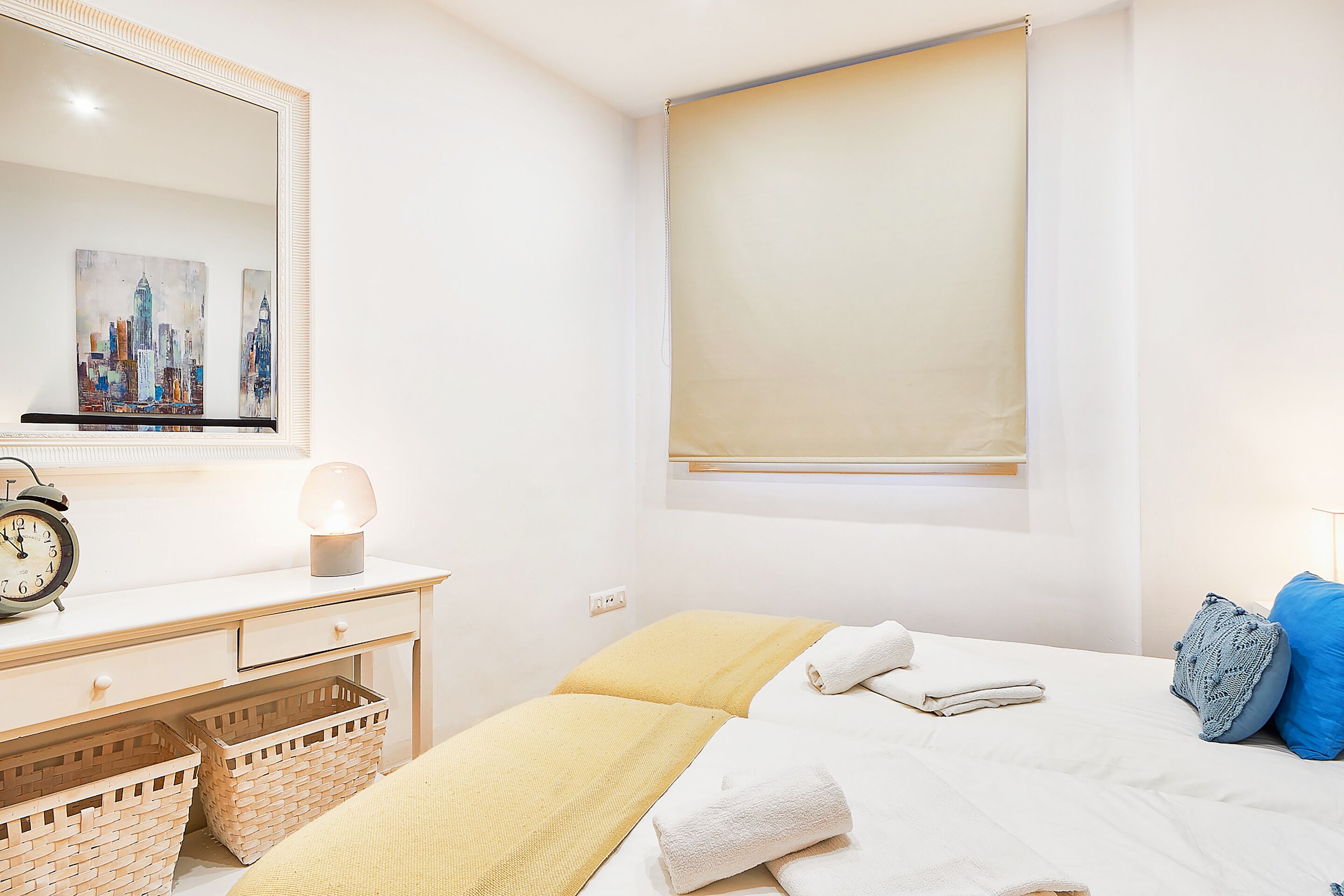 A hotel room with a bed, a desk with a lamp, a mirror, and a cityscape painting.