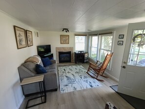 Een woonkamer met een bank, een televisie, een open haard, een schommelstoel en een groot raam.
