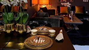 A dining area with a table set for four, featuring gold-rimmed plates and cutlery. There are two lamps with orange shades, and a large floral painting on the wall. - Dana Serviced Apartments2 (Riyadh)