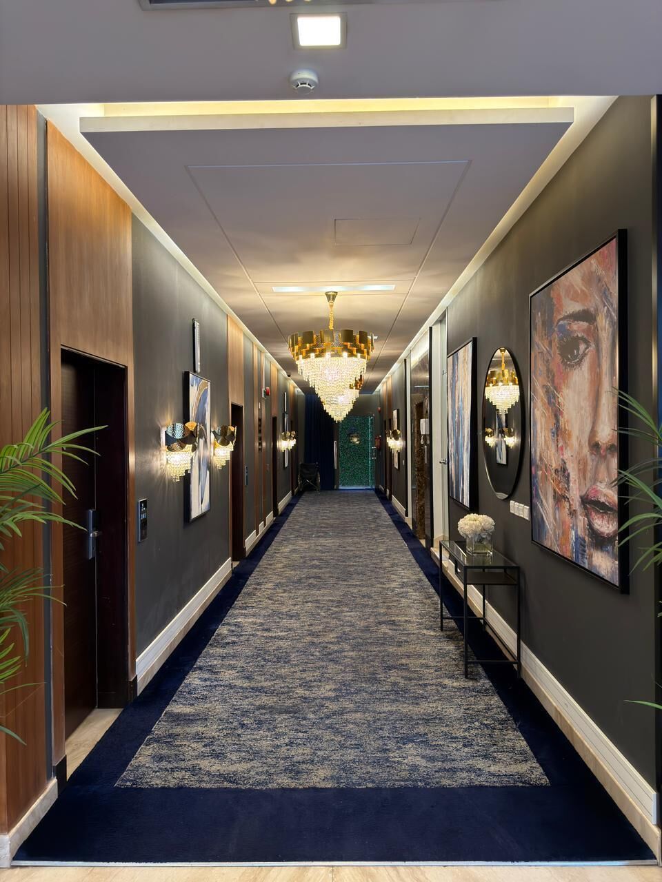 A hallway with a carpeted floor, wall-mounted light fixtures, and a chandelier.