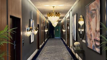 A hallway with a carpeted floor, wall-mounted light fixtures, and a chandelier.