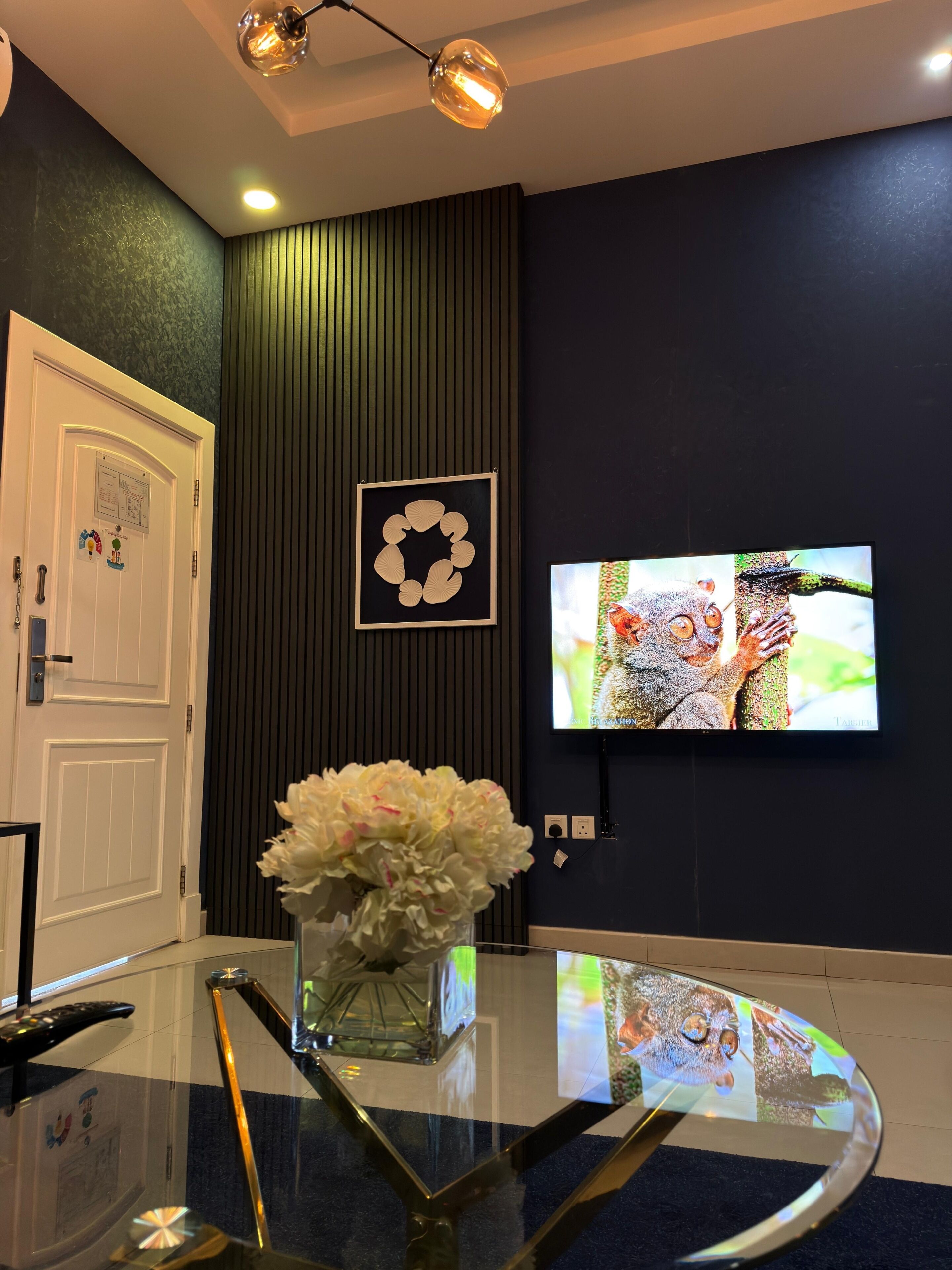 Una habitación con una televisión montada en la pared, una puerta blanca con un tablón de anuncios y una mesa de vidrio con un florero.