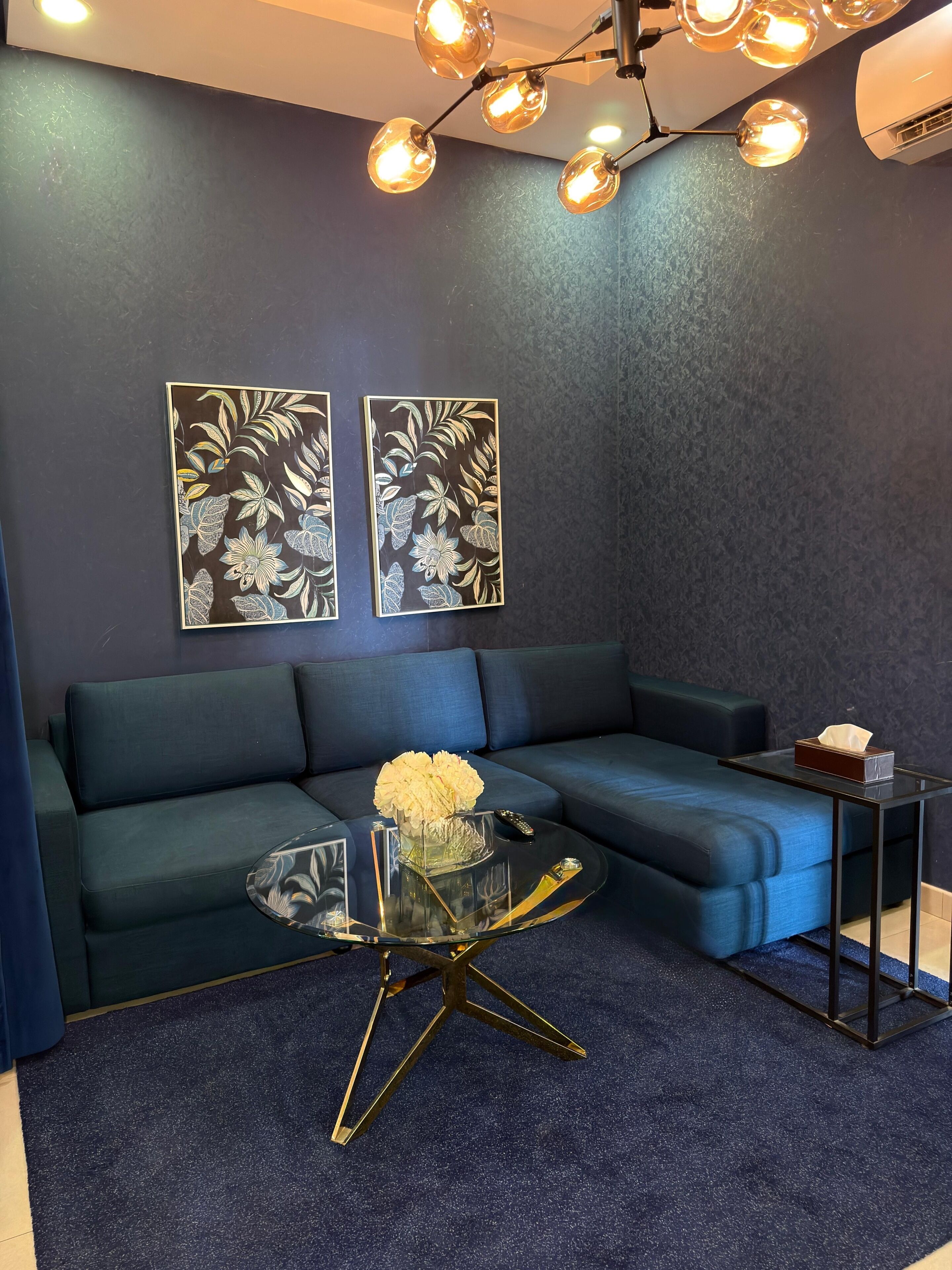 A modern living room with a sectional sofa, a glass coffee table with a gold base, and two wall-mounted artworks.