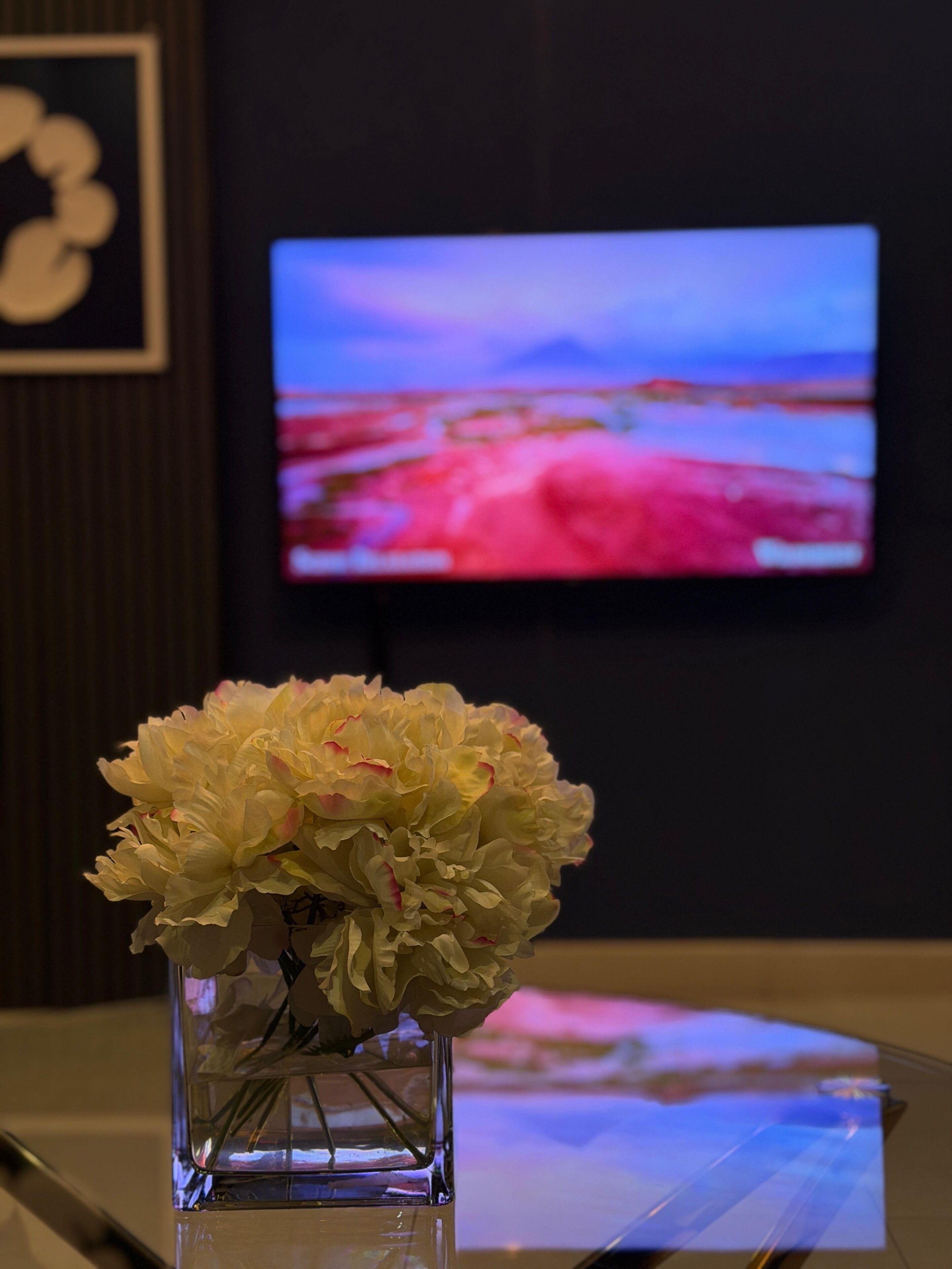 Um vaso com flores sobre uma superfície refletora, um quadro na parede e uma televisão exibindo uma paisagem.
