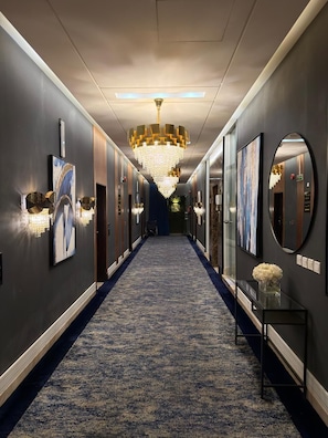 A hotel hallway with a blue carpet, chandeliers, framed artwork, and a mirror. - Dana Serviced Apartments2 (Riyadh)