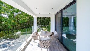 Un espace de restauration extérieur moderne, doté d’une balustrade en verre, d’une table ronde avec des chaises et offrant une vue sur une végétation luxuriante.