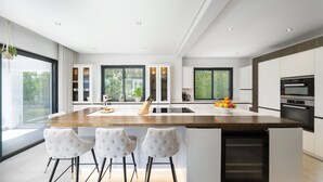 Une cuisine moderne dotée d’un grand îlot central, de tabourets de bar blancs et offrant une vue sur la verdure environnante.