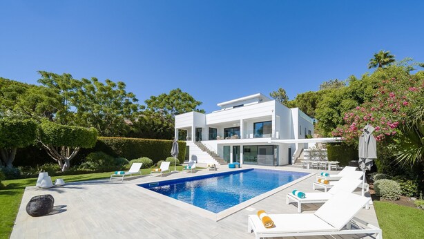 Une maison moderne à deux étages, dotée d’une piscine, d’un espace de détente extérieur et d’une végétation luxuriante.