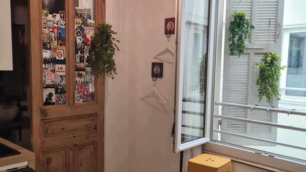 A room with a wooden door adorned with various stickers, a hanging light fixture, a window with shutters, and a stool. - La Sainte Madeleine (Gap)