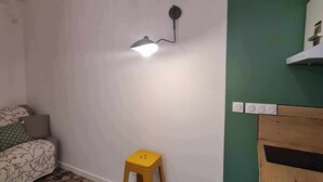 A room with a patterned floor, a yellow stool, a wall-mounted lamp, and a green wall with a shelf. - La Sainte Madeleine (Gap)
