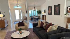 Ein geräumiges Wohnzimmer mit einem dunkelgrauen Sofa, einem runden Couchtisch und einem beige farbenen Sessel. Im Hintergrund ist eine Essecke mit einer Pendelleuchte und einer Pflanze zu sehen.