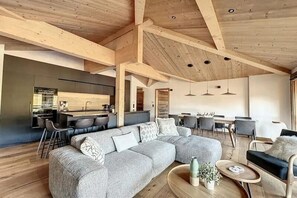 Un vaste espace de vie comprenant un grand canapé d’angle gris, une table basse ronde en bois et une cuisine à l’arrière-plan.
