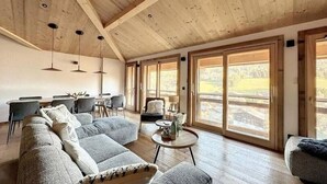 Tilava olohuone, jossa on puinen katto, suuret ikkunat, sohva, sohvapöytä ja ruokailutila tuoleineen.