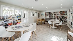 Une pièce spacieuse avec une table ronde, des chaises blanches, un baby-foot et des étagères remplies de livres.