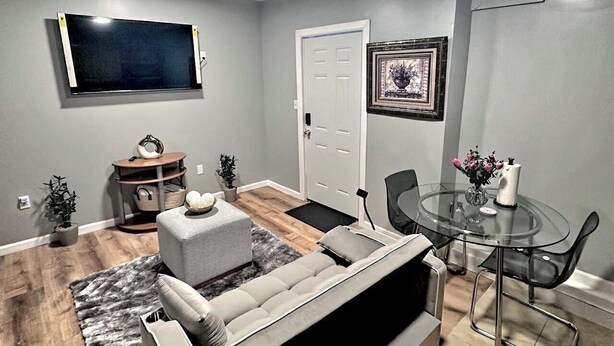 A modern living room with a grey sofa, a glass dining table, and a flat-screen TV mounted on the wall.
