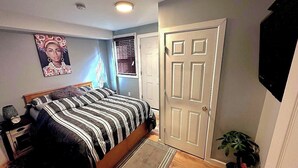 A bedroom with a bed, a nightstand, a wall-mounted TV, a door, and a window with blinds.