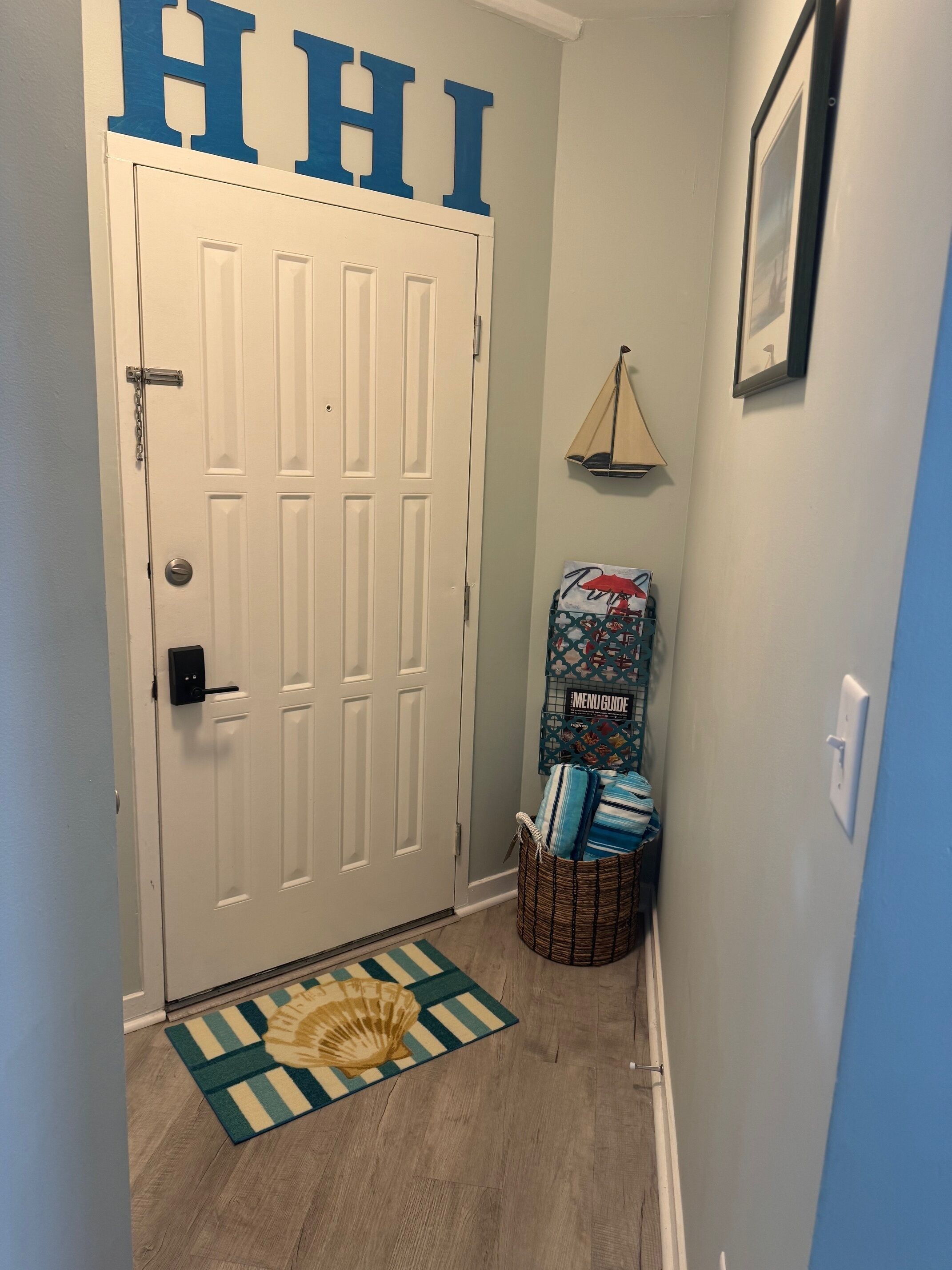 Un couloir avec une porte blanche, un paillasson orné d’un motif de coquillage, un panier rempli de coussins et un mur décoré d’une image de voilier.