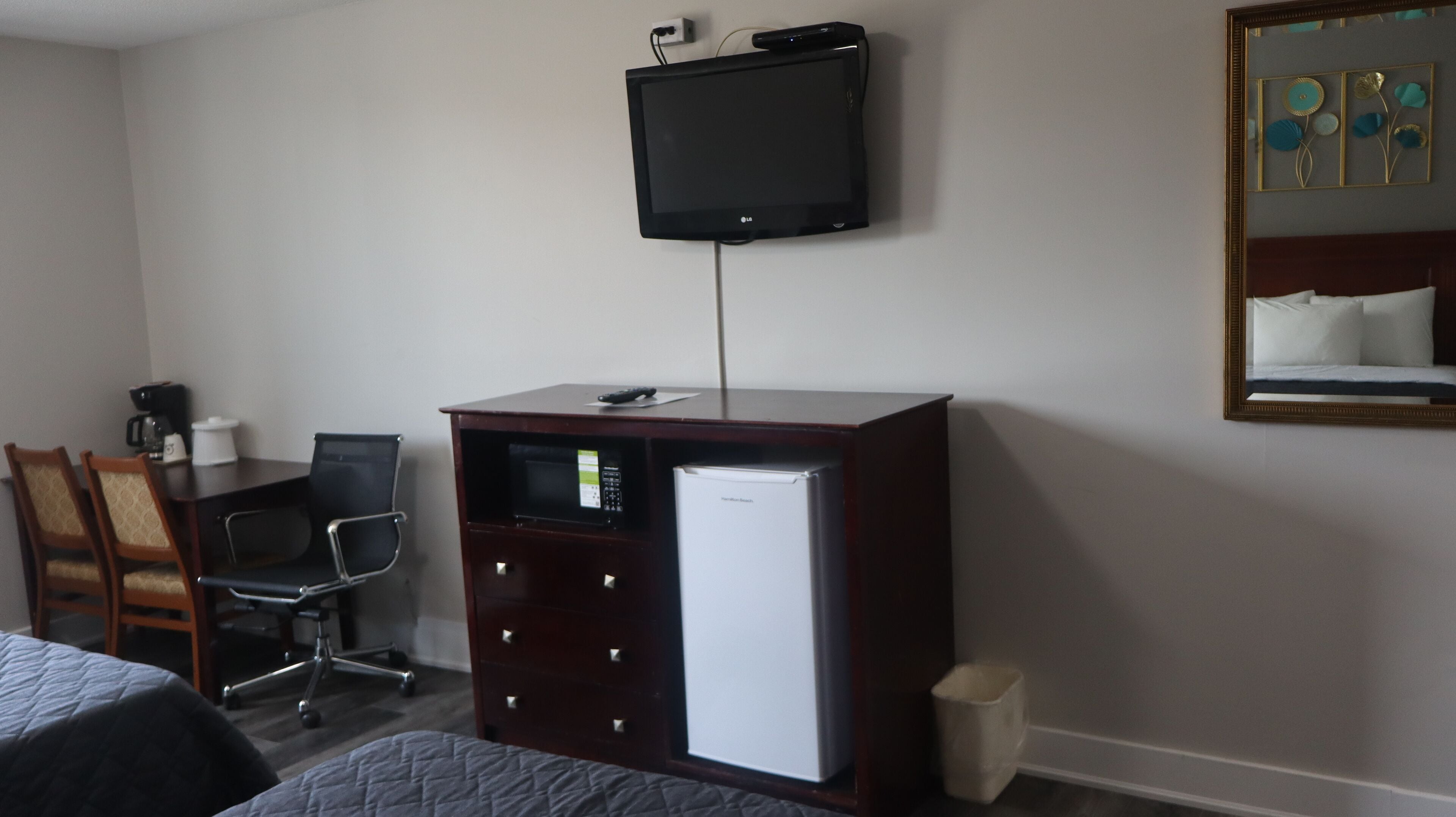 Una habitación de hotel con una cama, un escritorio con computadora, un refrigerador, un cesto de basura y un televisor montado en la pared.
