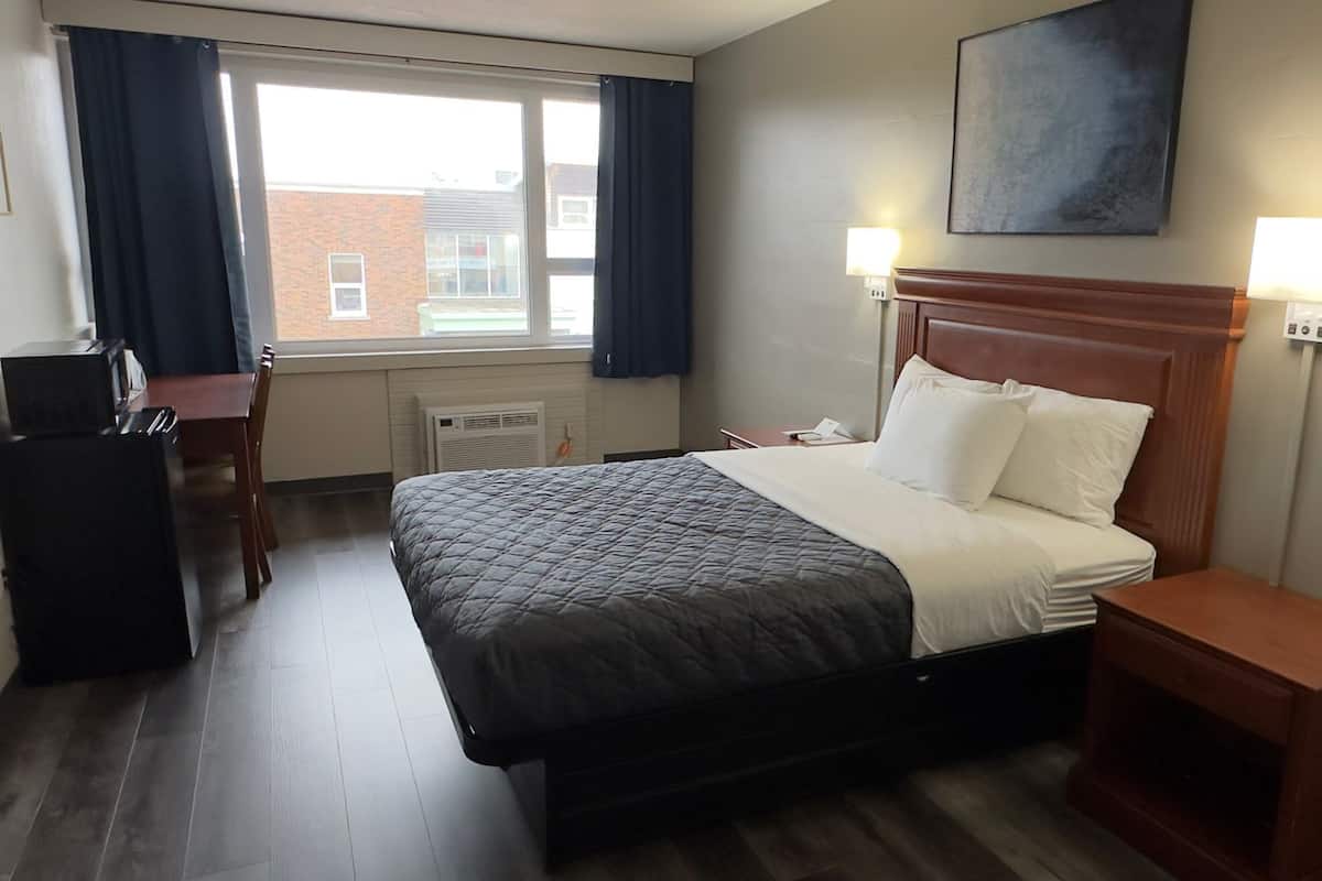 Habitación de hotel con cama, mesitas de noche, escritorio, silla, ventana con cortinas y una obra de arte colgada en la pared.
