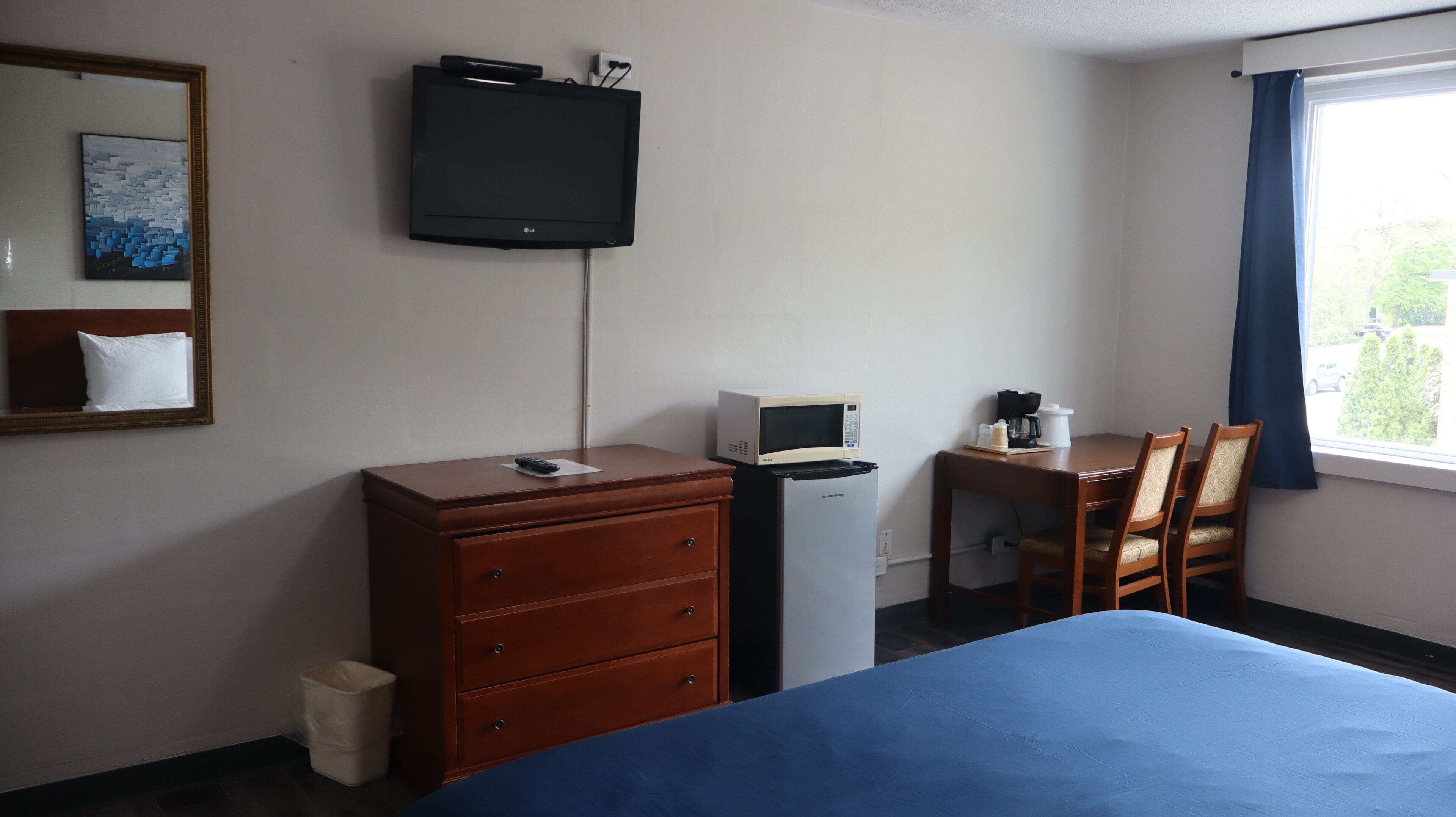 Quarto de hotel com cama, penteadeira, escrivaninha com micro-ondas, televisão e janela com cortinas.