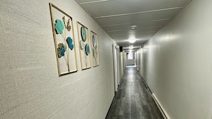 Um corredor comprido e estreito com piso de madeira e paredes cinza claro. Três obras de arte emolduradas na parede esquerda.