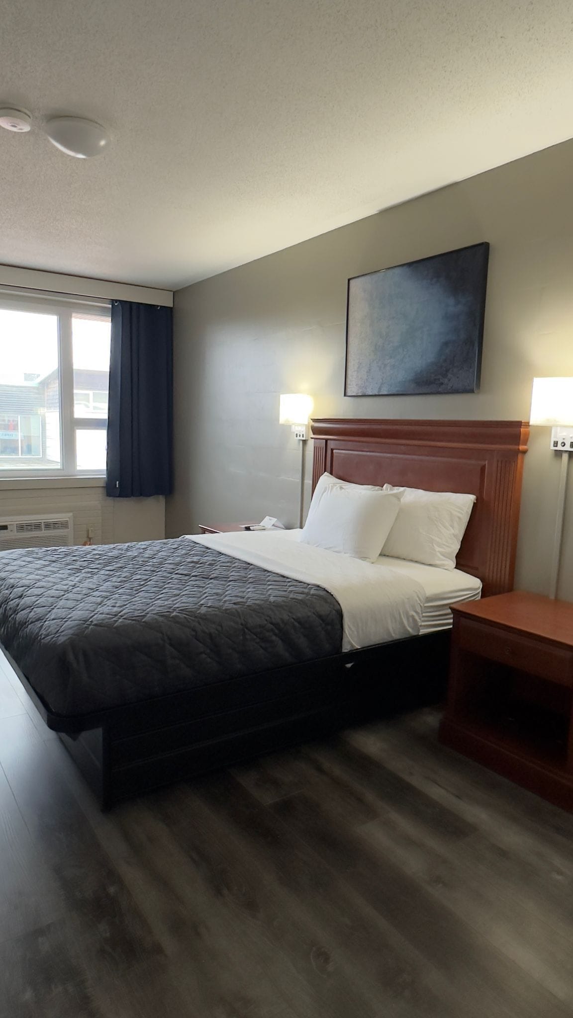 A hotel room with a bed, bedside tables, a headboard, a window with curtains, and a wall-mounted artwork.