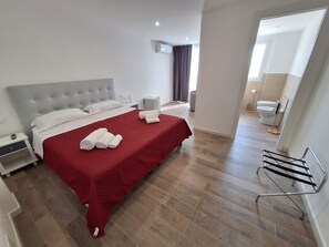 A hotel room with a large bed, a red bedspread, white pillows, a bedside table with a lamp, and a small nightstand.