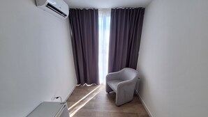 A small, modern room with a grey armchair, a white mini-fridge, and a window with dark curtains. - Room-double Bedroom With Double bed (Terrasini)