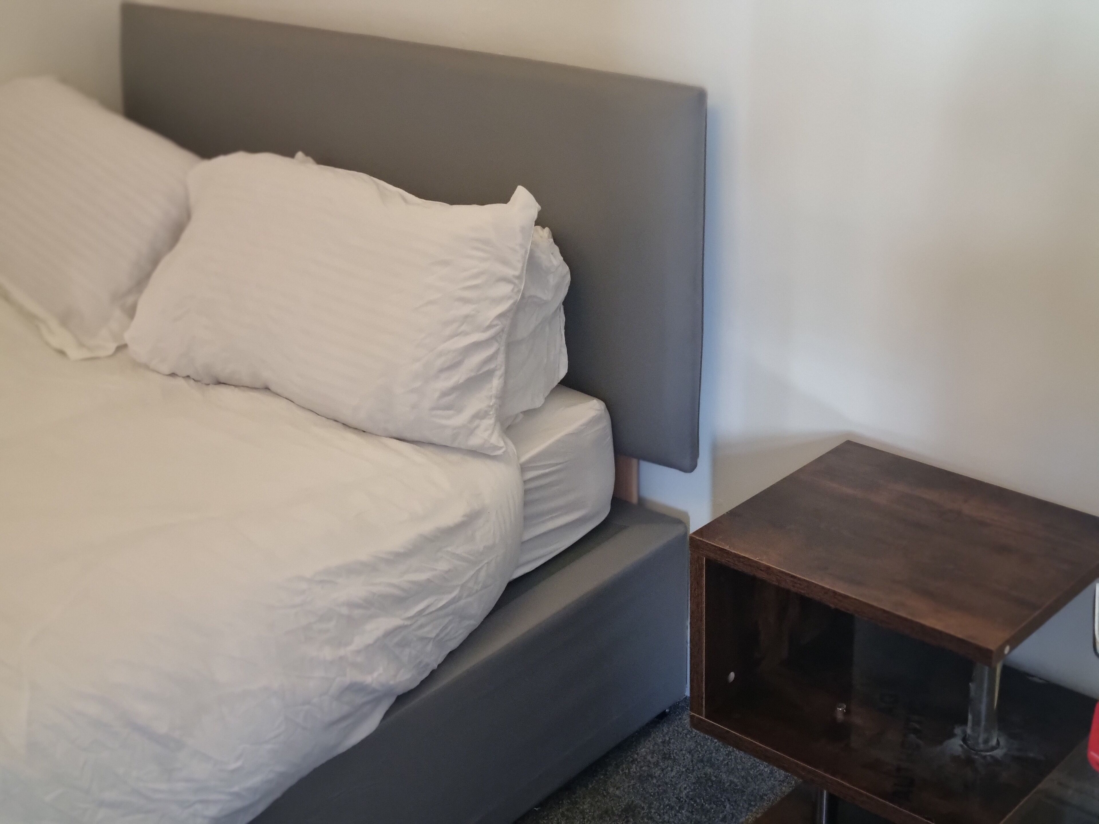 A bed with white bedding and a grey headboard, a small wooden nightstand with a glass top, and a grey carpeted floor.