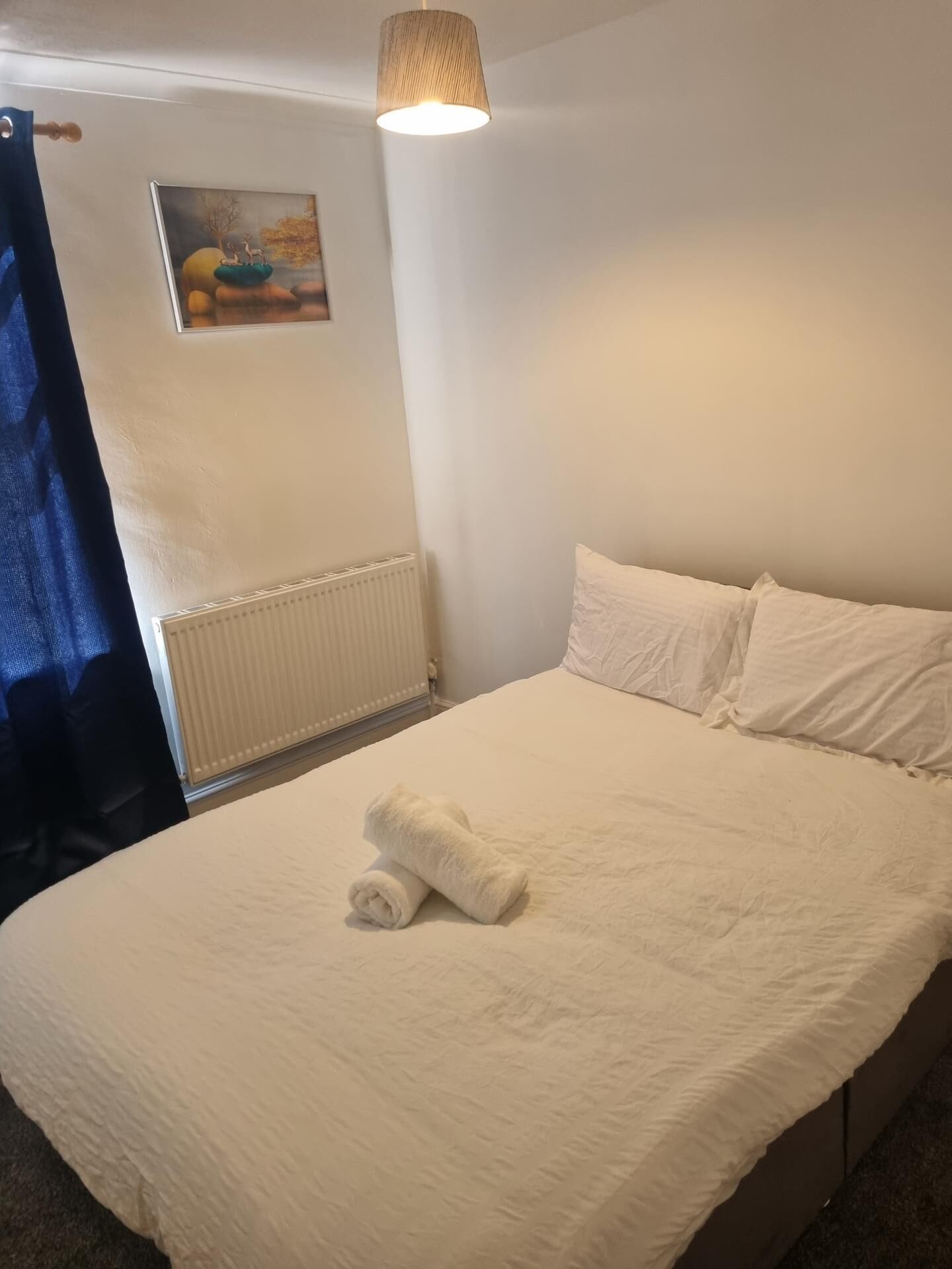 A neatly made bed with white linens and a rolled towel, a white radiator, and a painting on the wall.