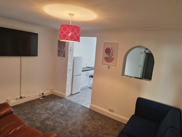 Una sala de estar con un sofá de cuero marrón, un sillón oscuro, una lámpara colgante roja con diseño, un televisor de pantalla plana montado en la pared y una cocina visible a través de un vano abierto.
