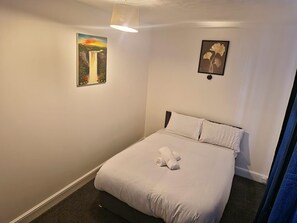 Una cama bien hecha con sábanas blancas y toallas enrolladas, un cuadro de una cascada en la pared izquierda y una obra de arte enmarcada con flores en la pared derecha.