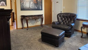 Una sala de estar con un sillón de cuero oscuro, un otoman a juego, una mesita consola de madera y un cuadro de un paisaje forestal.