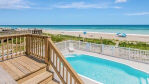 Una terraza de madera con piscina, con vista a una playa con sombrillas y un cielo despejado.