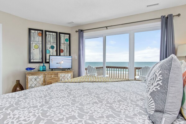 A bedroom with a large bed, a television, and a view of the ocean through a sliding glass door.