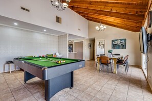 A pool table in a room with a dining area, wooden ceiling beams, and a TV mounted on the wall.