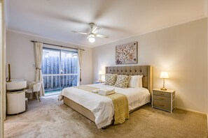 Une chambre à coucher comprenant un lit, des tables de chevet, un bureau, une chaise, un ventilateur de plafond et une fenêtre avec des rideaux.