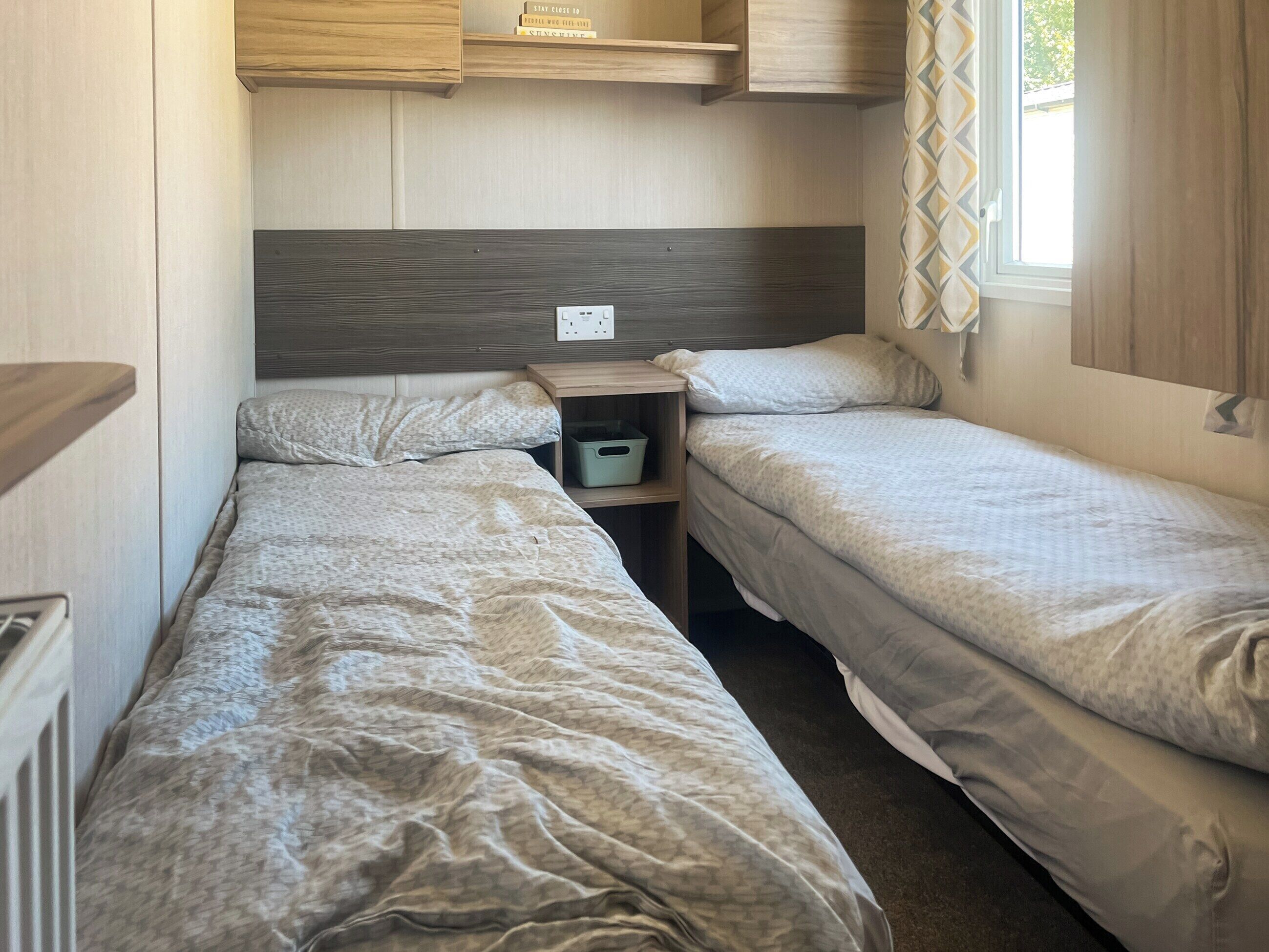 A compact RV bedroom with two single beds, a small wooden nightstand, and a shelf above the beds.