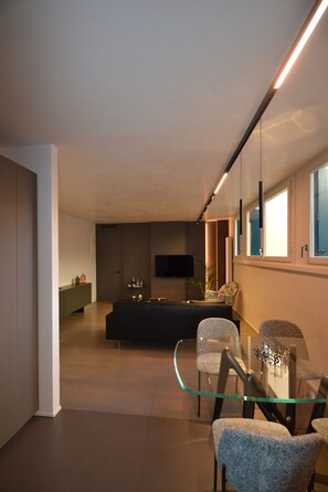 A modern living room with a glass coffee table, two upholstered chairs, a dark sofa, and a flat-screen TV mounted on the wall. - Nuovo Fiore (Pesaro)