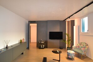 Ein modernes Wohnzimmer mit einem wandmontierten Flachbildfernseher, einem kleinen runden Tisch, einem blumengemusterten Sessel und einer Vitrine mit Dekorationsobjekten.