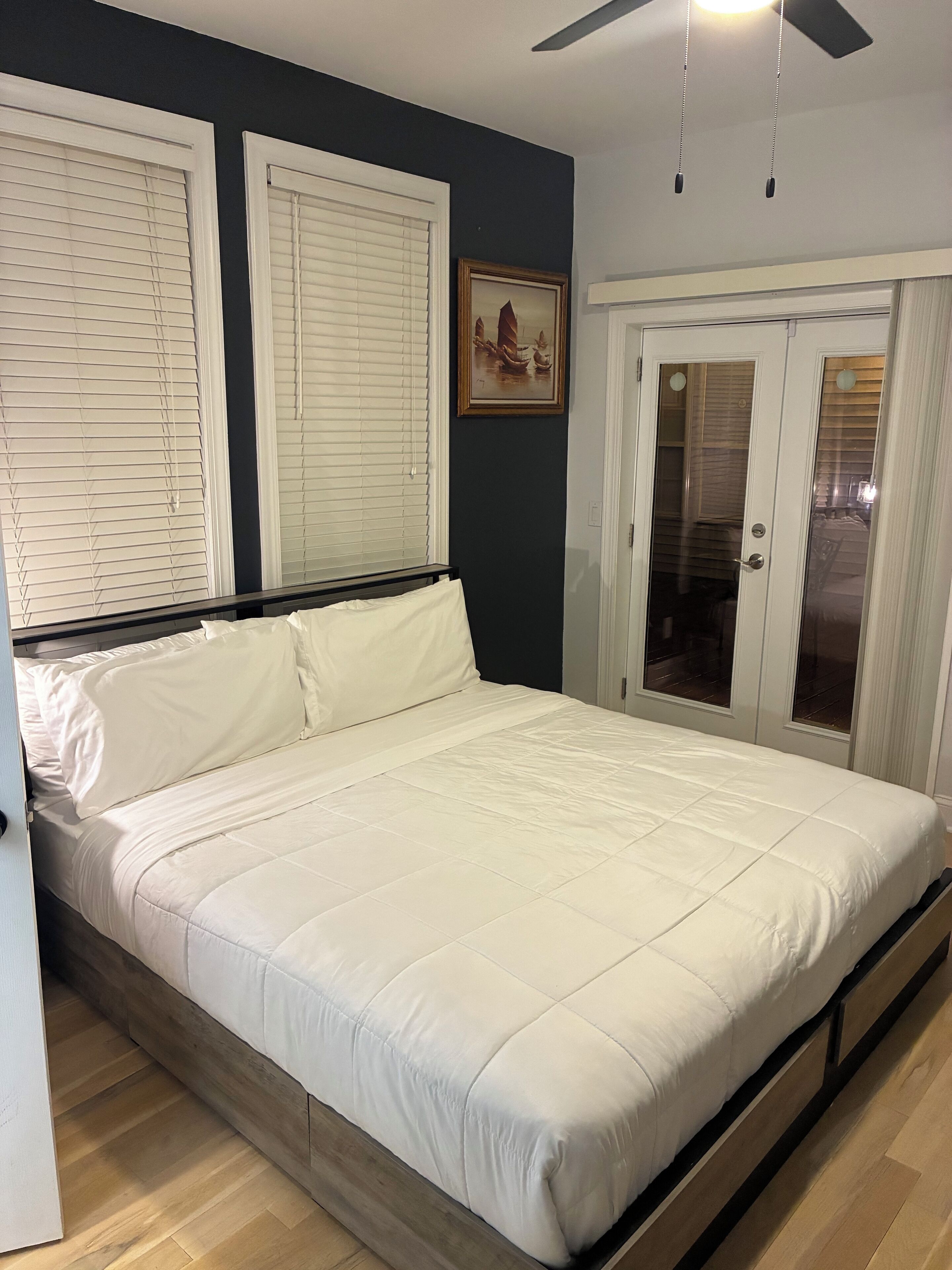 Ein Schlafzimmer mit einem Bett, zwei Fenstern mit Jalousien, einem Deckenventilator und einer Tür mit Glasfenster.