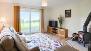 A living room with a sofa, TV, fireplace, and a view of a green landscape through the sliding doors. - The Lodge at Harvest Farm (Ludlow)