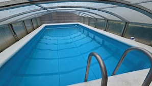 A swimming pool with a clear blue water surface, surrounded by a concrete deck and a glass ceiling structure.