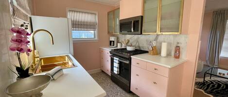 A compact kitchen with a microwave, stove, and sink. There is a window with blinds, a small rug, and a bowl on the counter.